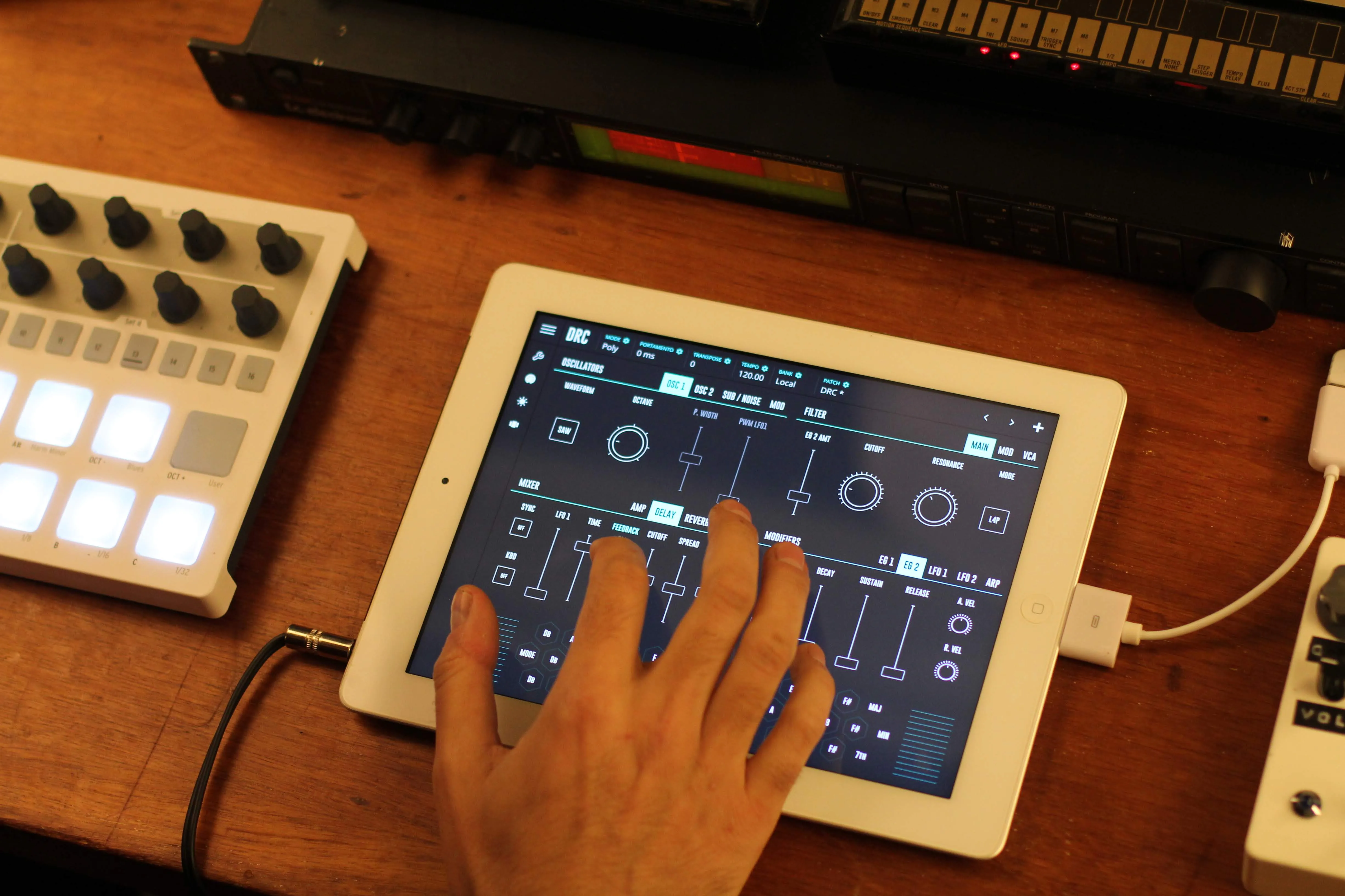 木製のデスクの上、白い iPad で DRC シンセのインターフェースを演奏する手。 左には小さなバックライト付き MIDI コントローラー、後ろにはラックマウントの ハードウェアユニット。