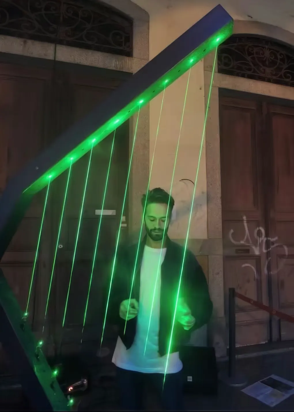 白い T シャツに濃い色のジャケットを着た男性が、ポルトガルの石畳の通りで Harpa Laser を 演奏している。背後の古い建物の木製の扉を、レーザーが鮮やかな緑の線で切り裂いている。